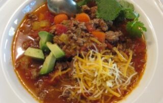 A bowl of chili with meat, cheese and avocado.